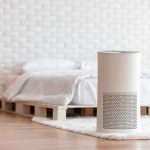 Un purificatore d'aria bianco in primo piano in una camera da letto con pavimento in parquet, pareti bianche con mattoni a vista e letto con struttura in pallet