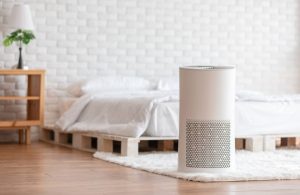 Un purificatore d'aria bianco in primo piano in una camera da letto con pavimento in parquet, pareti bianche con mattoni a vista e letto con struttura in pallet
