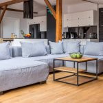Ampio loft con travi a vista e colonne in legno antico, parquet, salotto con divano grigio moderno e, sullo sfondo, una cucina