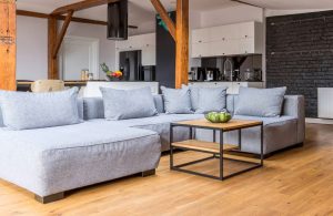 Ampio loft con travi a vista e colonne in legno antico, parquet, salotto con divano grigio moderno e, sullo sfondo, una cucina