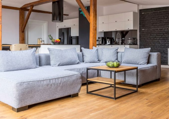 Ampio loft con travi a vista e colonne in legno antico, parquet, salotto con divano grigio moderno e, sullo sfondo, una cucina