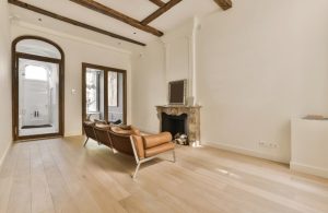 Salotto semivuoto con antico camino, pareti bianche decorate e con modanature, travi a vista scure, parquet chiaro, e divano vintage modernista in pelle