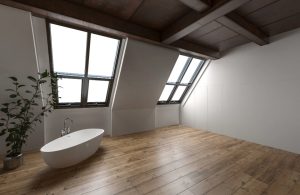 Sottotetto mansardato con finestre, travi a vista e soffitto a cassettoni in legno scuro, parquet medio-chiaro e vasca da bagno minimale bianca