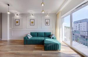 Salotto elegante in stile classico moderno con sofà a isola verde petrolio, pareti bianche con cornici, grande vetrata che si affaccia sulla città e pavimento in parquet
