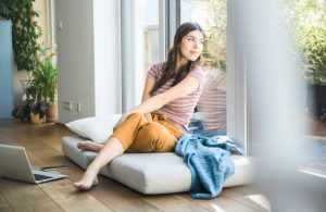 Una ragazza a piedi scalzi sul parquet, seduta su dei cuscini accanto a un laptop, guarda fuori da una grande finestra