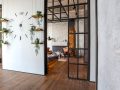 Ingresso di una casa in stile industrial minimale, con tramezzo bianco e porte a vetri nere, grande orologio sulla parete e piccole mensole con piantine. Si intravede un'altro ambiente: salotto con pareti in cemento grezzo, camino, poltrone e legna da ardere impilata. Il pavimento è in parquet scuro
