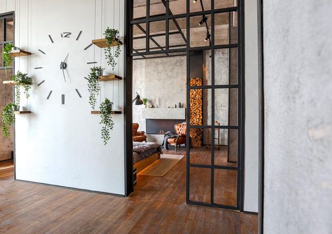 Ingresso di una casa in stile industrial minimale, con tramezzo bianco e porte a vetri nere, grande orologio sulla parete e piccole mensole con piantine. Si intravede un'altro ambiente: salotto con pareti in cemento grezzo, camino, poltrone e legna da ardere impilata. Il pavimento è in parquet scuro