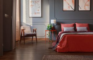 Camera da letto con pareti in due diversi toni di grigio e cornici decorative. Ci sono stampe astratte appese ai muri. Il letto è rosso e grigio, accanto c'è un tavolino arancione con sopra una lampada e una pianta. Vicino alla finestra c'è una poltrona con struttura in legno. Il pavimento è in parquet scuro