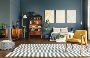 Grande camera da letto con pareti blu-grigio, pavimento in parquet scuro, letto matrimoniale sui toni del bianco e del blu polvere, poltrona in pelle giallo senape e struttura in legno, mobili vintage in legno scuro, tappeto con motivo geometrico bianco e grigio