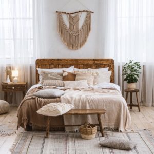 Camera da letto in stile boho-chic con pareti bianche. Letto in legno con testata semplice e numerosi cuscini. Pavimento in legno chiaro. Presenti piante da interno, un tappeto intrecciato e tende trasparenti. Mobili in legno chiaro e tessuti naturali.