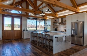 Cucina ampia e luminosa, caratterizzata da un soffitto a volta con travi in legno a vista. Il pavimento è in legno scuro, a listoni, con effetto invecchiato, mentre i pensili e il top della cucina sono bianchi. Al centro della stanza è presente un'isola con piano in marmo bianco e sgabelli in legno. Una finestra panoramica e una porta a vetri offrono una vista sull'esterno