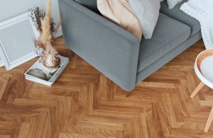 Una porzione di un soggiorno moderno. Il pavimento è caratterizzato da un parquet a spina di pesce in tonalità calde. Un divano rivestito in tessuto grigio chiaro occupa la parte centrale dell'inquadratura. Sul divano sono presenti diversi cuscini decorativi. A fianco del divano, un tavolino basso con gambe in legno e piano rotondo. Sulla superficie del tavolino è appoggiato un oggetto decorativo. In primo piano, su un piccolo tavolino, sono visibili libri e un vaso contenente rami secchi.