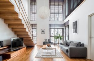 Soggiorno con soffitto a doppia altezza e grandi finestre. Presente una scala in legno che collega i due piani. Il pavimento è in legno di tonalità calda. L'arredamento include un divano grigio, un tappeto chiaro, un tavolino da caffè e piante verdi