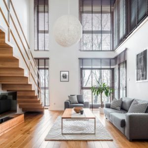 Soggiorno con soffitto a doppia altezza e grandi finestre. Presente una scala in legno che collega i due piani. Il pavimento è in legno di tonalità calda. L'arredamento include un divano grigio, un tappeto chiaro, un tavolino da caffè e piante verdi