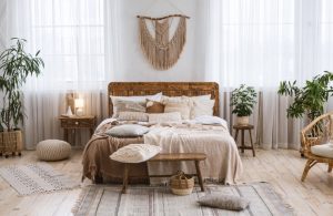 Camera da letto in stile boho-chic con pareti bianche. Letto in legno con testata semplice e numerosi cuscini. Pavimento in legno chiaro. Presenti piante da interno, un tappeto intrecciato e tende trasparenti. Mobili in legno chiaro e tessuti naturali.