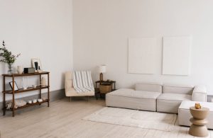 Soggiorno caratterizzato da pareti bianche e un pavimento in legno chiaro. Al centro della stanza è presente un divano componibile di colore chiaro, affiancato da un tavolino basso con piano in legno. In un angolo, una poltrona imbottita è completata da un plaid a righe. Una libreria in legno ospita oggetti decorativi e libri. Due quadri dalle linee essenziali adornano la parete.