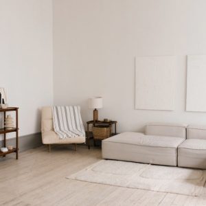 Soggiorno caratterizzato da pareti bianche e un pavimento in legno chiaro. Al centro della stanza è presente un divano componibile di colore chiaro, affiancato da un tavolino basso con piano in legno. In un angolo, una poltrona imbottita è completata da un plaid a righe. Una libreria in legno ospita oggetti decorativi e libri. Due quadri dalle linee essenziali adornano la parete.