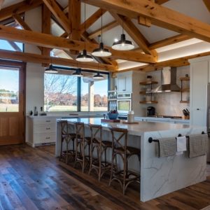 Cucina ampia e luminosa, caratterizzata da un soffitto a volta con travi in legno a vista. Il pavimento è in legno scuro, a listoni, con effetto invecchiato, mentre i pensili e il top della cucina sono bianchi. Al centro della stanza è presente un'isola con piano in marmo bianco e sgabelli in legno. Una finestra panoramica e una porta a vetri offrono una vista sull'esterno