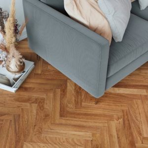 Una porzione di un soggiorno moderno. Il pavimento è caratterizzato da un parquet a spina di pesce in tonalità calde. Un divano rivestito in tessuto grigio chiaro occupa la parte centrale dell'inquadratura. Sul divano sono presenti diversi cuscini decorativi. A fianco del divano, un tavolino basso con gambe in legno e piano rotondo. Sulla superficie del tavolino è appoggiato un oggetto decorativo. In primo piano, su un piccolo tavolino, sono visibili libri e un vaso contenente rami secchi.